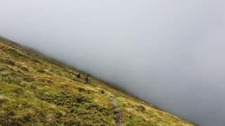 Two hikers on a grassy slope of Mali i Livadhit on Oshlak as fog swallows the valley beside them